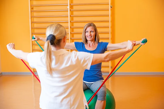 Die Patientin und die Therapeutin führen eine Übung am Gymnastikball aus, man sieht die Therapeutin nur von hinten