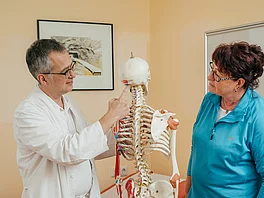 Patientin mit Morbus Bechterew bespricht sich mit einem Arzt im Gasteiner Heilstollen, der Arzt zeigt am Skelett etwas vor.