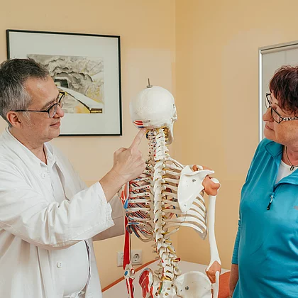 Patientin mit Morbus Bechterew bespricht sich mit einem Arzt im Gasteiner Heilstollen, der Arzt zeigt am Skelett etwas vor.