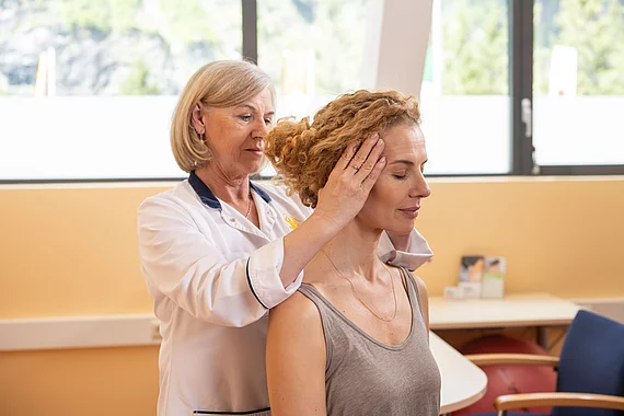 Eine Ärztin führt bei einer sitzenden Patientin eine manuelle Untersuchung der Halswirbelsäule durch. Sie hält den Kopf der Patientin seitlich, um die Beweglichkeit zu testen – Teil der Diagnostik bei Morbus Bechterew im Gasteiner Heilstollen.