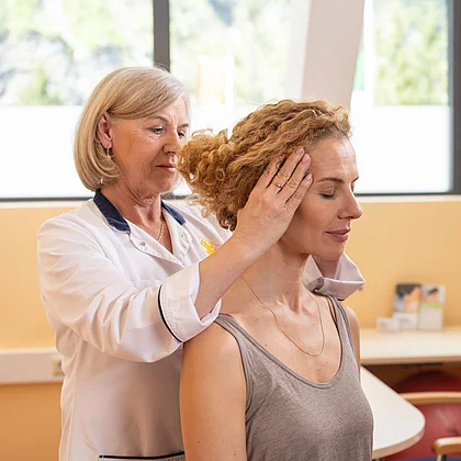 Eine Ärztin führt bei einer sitzenden Patientin eine manuelle Untersuchung der Halswirbelsäule durch. Sie hält den Kopf der Patientin seitlich, um die Beweglichkeit zu testen – Teil der Diagnostik bei Morbus Bechterew im Gasteiner Heilstollen.