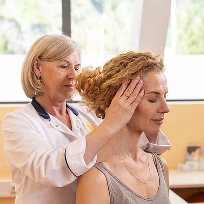 Eine Ärztin führt bei einer sitzenden Patientin eine manuelle Untersuchung der Halswirbelsäule durch. Sie hält den Kopf der Patientin seitlich, um die Beweglichkeit zu testen – Teil der Diagnostik bei Morbus Bechterew im Gasteiner Heilstollen.