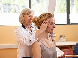 Eine Ärztin führt bei einer sitzenden Patientin eine manuelle Untersuchung der Halswirbelsäule durch. Sie hält den Kopf der Patientin seitlich, um die Beweglichkeit zu testen – Teil der Diagnostik bei Morbus Bechterew im Gasteiner Heilstollen.