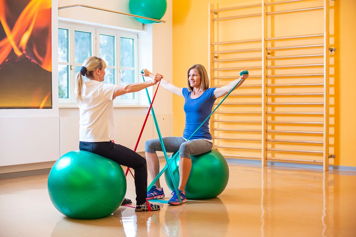 Die Therapeutin und die Patientin machen sitzend eine Übung am Gymnastikball mit elastischen Sportbändern im Gasteiner Heilstollen