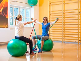 Die Therapeutin und die Patientin machen sitzend eine Übung am Gymnastikball mit elastischen Sportbändern im Gasteiner Heilstollen